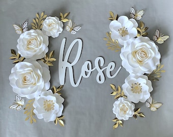 May include: White paper flowers with gold accents and butterflies, arranged around the word "Rose" in white letters.
