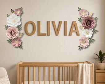 May include: Wooden letters spelling "OLIVIA" are mounted on a wall, flanked by paper flower arrangements in white, pink, and mauve. A wooden crib is visible below, suggesting a nursery theme. The flowers have gold and green accents.