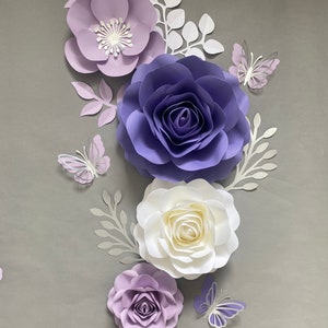 May include: A collection of handcrafted paper flowers in shades of light purple, deep violet, and white, arranged with white paper leaves and three light purple paper butterflies. The flowers feature layered petals and a central white stamen detail.