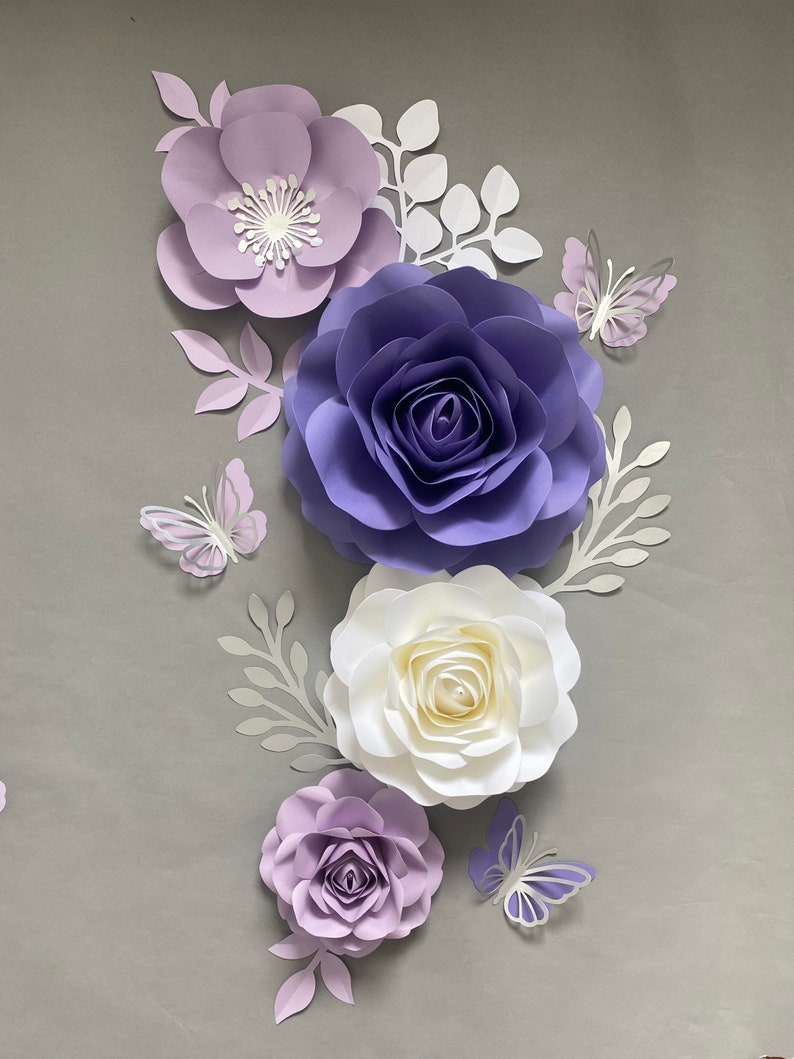 May include: A collection of handcrafted paper flowers in shades of light purple, deep violet, and white, arranged with white paper leaves and three light purple paper butterflies. The flowers feature layered petals and a central white stamen detail.