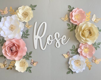May include: A decorative wall art piece featuring paper flowers in white, blush pink, and cream, with gold accents and butterflies. The word "Rose" is displayed in white script, creating a floral and whimsical design.