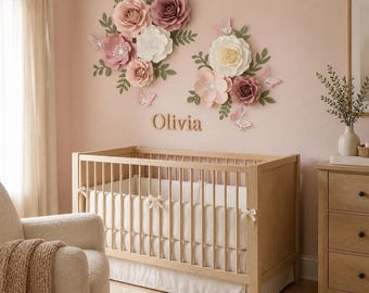 May include: A wooden crib with white bedding sits below a wall display of large paper flowers in shades of pink, cream, and mauve, accented with green leaves and small butterflies. The name Olivia is mounted on the wall in wooden script lettering.