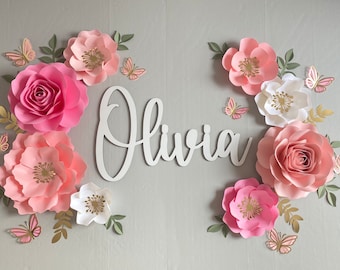 May include: A white wooden sign with the name "Olivia" written in cursive script. The sign is surrounded by pink and white paper flowers and gold butterflies.
