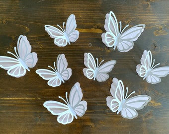 May include: Seven white paper butterflies with a light pink overlay are arranged on a dark brown wooden surface. The butterflies are all facing the same direction and are slightly overlapping each other.
