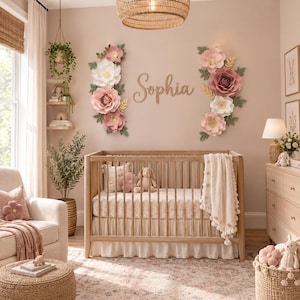 May include: A wooden baby crib with a white ruffled skirt and a cream-colored knit blanket. Above the crib, the name Sophia is written in wooden script letters, flanked by two large arrangements of pink and white paper flowers.
