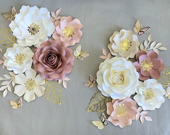 May include: Arrangement of paper flowers in white, blush pink, and mauve, with gold accents and butterfly cutouts. The floral display includes various flower types and leaf details, set against a gray backdrop.