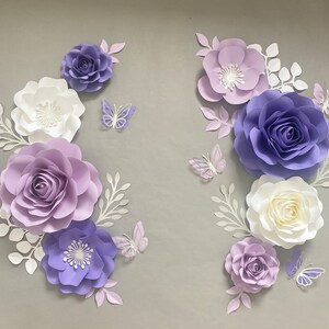 May include: A collection of paper flowers and butterflies in shades of purple, lavender, and white. The arrangement includes various sizes of layered roses and blossoms, accompanied by delicate white paper leaves and intricate butterfly cutouts.