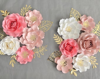 May include: A set of paper flowers in shades of pink and white with gold accents. The flowers are arranged in a cluster and are surrounded by gold leaves.
