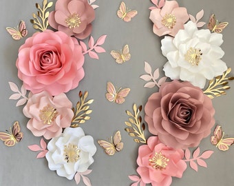 Puede incluir: Una colección de flores de papel rosa y blanco con detalles dorados. Las flores están dispuestas en un patrón circular con mariposas dispersas por todas partes.