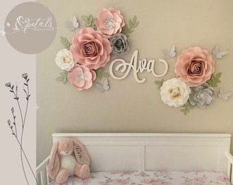 May include: A nursery wall decor featuring a large pink, white, and gray paper flower arrangement with the name "Ava" in white lettering. The flowers are arranged in a cluster with butterflies and greenery.
