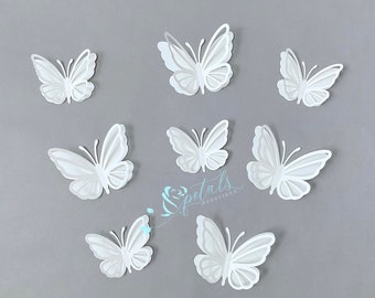 May include: Seven white paper butterflies of various sizes are arranged on a gray background. The butterflies are cut out and have a 3D effect.