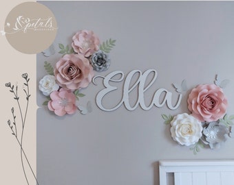 May include: A wall decoration featuring the name "Ella" in white script, surrounded by large paper flowers in shades of pink, white, and gray, with green leaves and butterfly accents. The flowers are arranged in clusters.