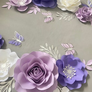 May include: A collection of handcrafted paper flowers in shades of lavender, deep purple, and white, arranged with intricate paper butterflies and delicate leafy sprigs. The set features various bloom styles, including layered roses and flat-petaled flowers.