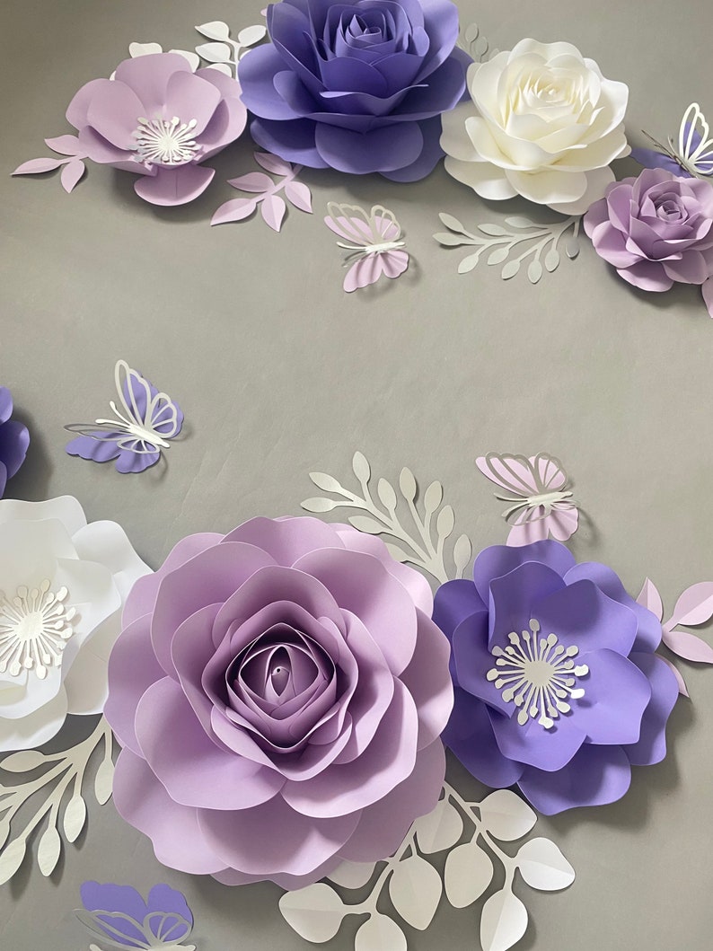 May include: A collection of handcrafted paper flowers in shades of lavender, deep purple, and white, arranged with intricate paper butterflies and delicate leafy sprigs. The set features various bloom styles, including layered roses and flat-petaled flowers.