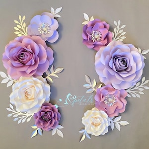 May include: A collection of handcrafted paper flowers in shades of lavender, purple, and cream, arranged with white paper leaves. The flowers feature layered petals and central accents. The text Petals Redefined is visible in the center.