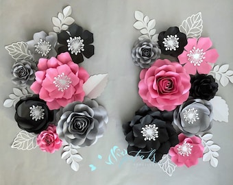 May include: Arrangement of paper flowers in pink, black, and gray, with white leaf accents. The flowers vary in size and style, creating a layered effect. The background is a light gray, providing a neutral backdrop for the colorful blooms.
