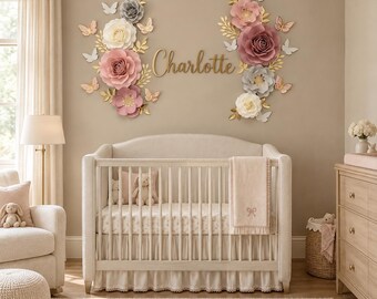 May include: A wall display featuring large paper flowers in shades of pink, white, and gray, accented with gold leaves and butterflies. The name Charlotte is written in gold script in the center, positioned above a cream-colored wooden crib.