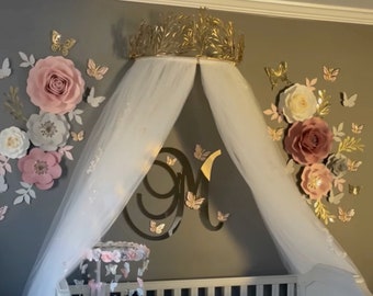 May include: A nursery scene with a white crib, a canopy, and wall decorations. The wall features large paper flowers in pink, white, and gray, along with gold butterflies and a gold crown. A mobile hangs above the crib.