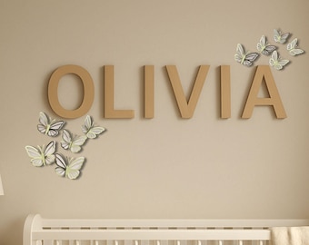 May include: Wooden letters spelling "OLIVIA" are mounted on a beige wall, accompanied by butterfly decorations. The butterflies have white, gray, and light green wings. A white crib is visible in the lower portion of the image.