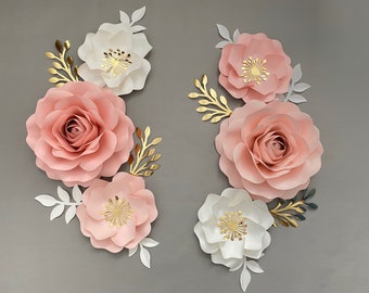 May include: Arrangement of large paper flowers in shades of pink and white, accented with gold and white leaf details. The flowers are arranged in a symmetrical pattern against a gray background, creating a decorative floral display.