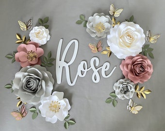 May include: A white paper sign with the word "Rose" in cursive script, surrounded by paper flowers in shades of pink, white, and gray. The flowers are decorated with gold glitter and butterflies.