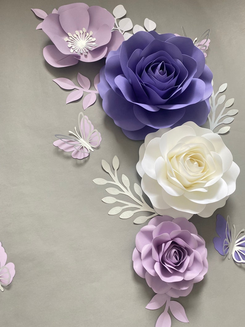 May include: A collection of handcrafted paper flowers and butterflies in shades of purple, lavender, and white. The arrangement includes large layered roses and smaller blossoms, accented by delicate paper leaves and butterfly cutouts.