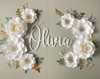 May include: A decorative wall art piece featuring the name "Olivia" in cursive script, surrounded by large white paper flowers, gold accents, and butterfly cutouts. The flowers have gold centers and are paired with green and gold leaves.