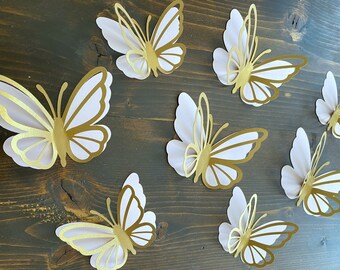 May include: Eight white paper butterflies with gold accents are arranged on a dark brown wooden surface. The butterflies are all facing the same direction and are evenly spaced apart.