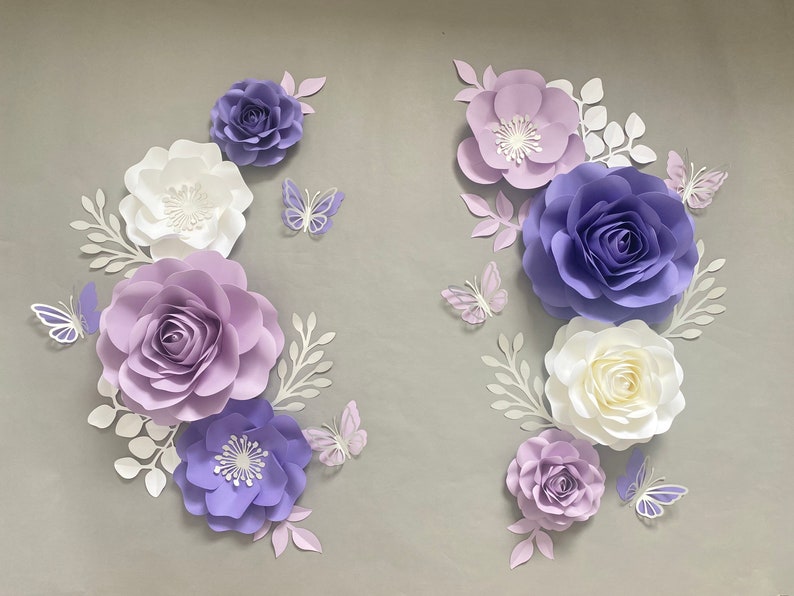 May include: A collection of paper flowers and butterflies in shades of purple, lavender, and white. The arrangement includes various sizes of layered roses and blossoms, accompanied by delicate white paper leaves and intricate butterfly cutouts.