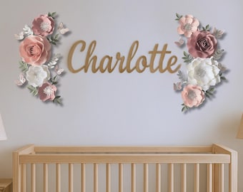 May include: A nursery wall decoration featuring the name "Charlotte" in wooden script, flanked by paper flowers in shades of pink, white, and mauve, with butterfly accents. A wooden crib is in the foreground.