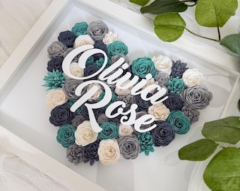 May include: A framed heart-shaped floral arrangement. The heart is composed of paper flowers in shades of gray, teal, and cream. The names "Olivia Rose" are displayed in white script across the center. The frame is white, and the background is a sheer white fabric.