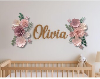 May include: A nursery wall decoration featuring the name "Olivia" in wooden script, flanked by paper flower arrangements in shades of pink, mauve, and white, with green foliage accents. A wooden crib and stuffed animal are in the foreground.