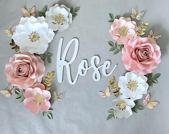 May include: A white background with a pink and white paper flower arrangement. The flowers are arranged in a circle around the word "Rose" in white letters. The flowers have gold centers and are accented with butterflies.