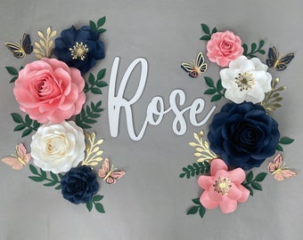 May include: A paper flower arrangement with the name "Rose" in white letters. The flowers are in shades of pink, white, and navy blue with gold accents. Butterflies are scattered throughout the arrangement.