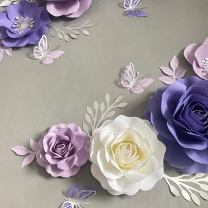May include: A collection of handcrafted paper flowers in shades of lavender, deep purple, and white, arranged with die-cut paper leaves and several small paper butterflies in matching purple and white tones, creating a layered floral display.