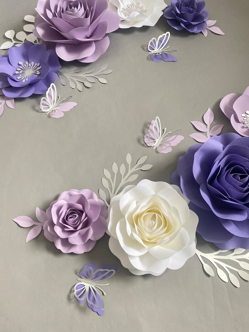 May include: A collection of handcrafted paper flowers in shades of lavender, deep purple, and white, arranged with die-cut paper leaves and several small paper butterflies in matching purple and white tones, creating a layered floral display.