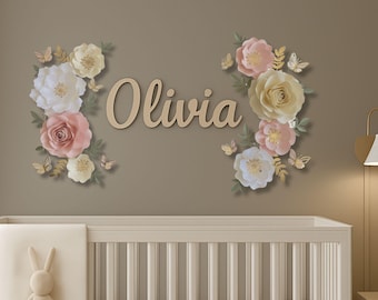 May include: A wooden name, "Olivia", is surrounded by paper flowers in white, pink, and yellow, with gold butterflies. The floral arrangement is mounted on a neutral-toned wall above a crib.