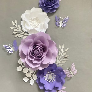 May include: A collection of paper flowers and butterflies in shades of purple, lavender, and white. The arrangement includes three large layered paper roses and two smaller open-petaled flowers, accented by white paper leaves and three butterflies.
