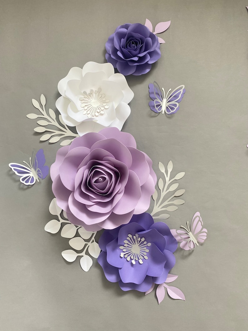May include: A collection of paper flowers and butterflies in shades of purple, lavender, and white. The arrangement includes three large layered paper roses and two smaller open-petaled flowers, accented by white paper leaves and three butterflies.