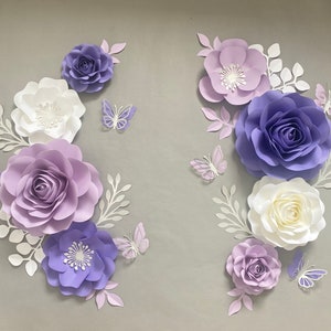 May include: A collection of paper flowers and butterflies in shades of purple, lavender, and white. The arrangement includes various sizes of layered roses and blossoms, accompanied by delicate white paper leaves and intricate butterfly cutouts.