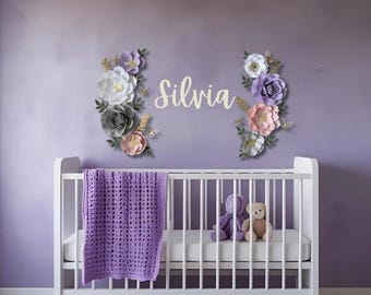 May include: A white crib with a purple blanket, two stuffed animals, and a floral wall decoration spelling "Silvia". The wall art features paper flowers in white, gray, pink, and purple, with gold accents, set against a lavender wall.