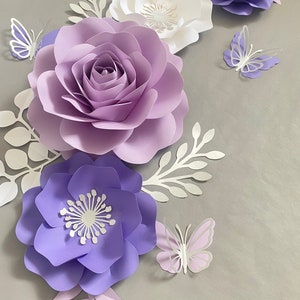 May include: A collection of handcrafted paper flowers in shades of lavender, purple, and white, accompanied by white paper foliage and three-dimensional paper butterflies in matching purple and white tones, arranged in a decorative floral display.