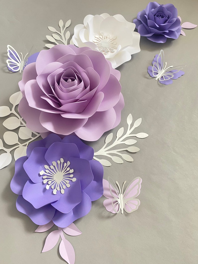 May include: A collection of handcrafted paper flowers in shades of lavender, purple, and white, accompanied by white paper foliage and three-dimensional paper butterflies in matching purple and white tones, arranged in a decorative floral display.