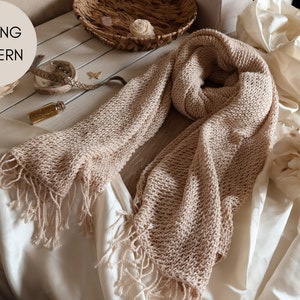 May include: A beige knitted scarf with fringe on the ends. The scarf is laid out on a white fabric surface.
