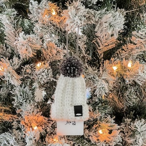 May include: A white knit beanie Christmas ornament with a dark gray pom-pom and a small black tag. A white rectangular tag with the word "Joy" is attached to the bottom of the beanie. The ornament hangs on a snow-covered Christmas tree.