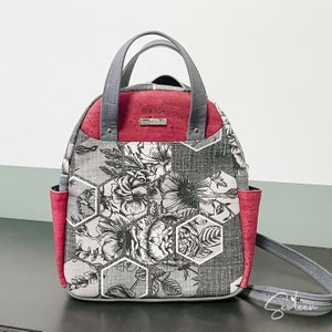 Peut inclure: Un sac à dos à motif floral gris et rouge avec un tissu ressemblant à du liège. Le sac a une poignée supérieure et des bretelles réglables.