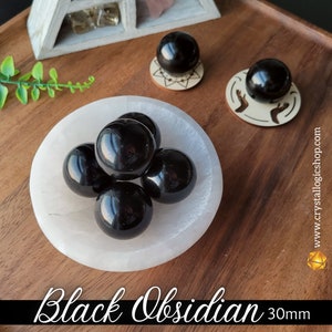 May include: Five black obsidian spheres, each measuring 30mm in diameter, are arranged in a white selenite bowl. The spheres are smooth and polished, reflecting light.