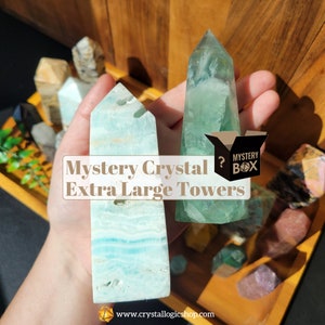 May include: Two large, extra-large crystal towers, one light blue and one green, are held in a hand. The towers are on a wooden surface with other smaller crystals. The text "Mystery Crystal Extra Large Towers" is on the image.
