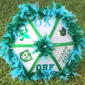 May include: A white graduation cap decorated with green and blue feathers. The cap has six triangular sections with green trim and various graphics, including Greek letters, a fleur-de-lis, and the text "CLASS OF 2023".