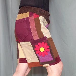 May include: Patchwork skirt featuring a variety of fabrics in shades of brown, beige, burgundy, and purple. The skirt has a floral applique on a pocket and an elastic waistband. The skirt is knee-length.